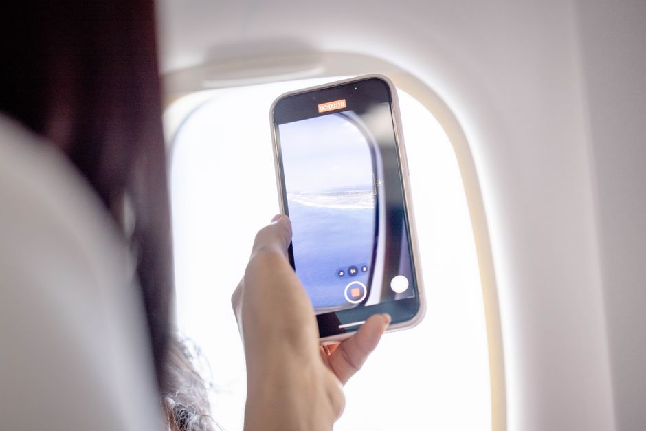 Woman filming out an airplane window using a smartphone, capturing a beautiful aerial view.