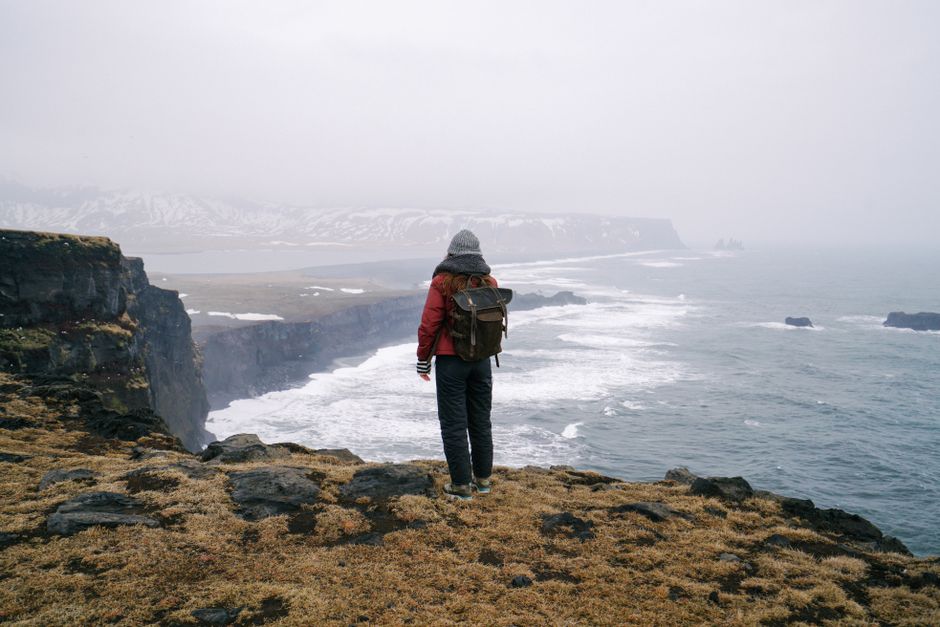 Person with backpack stands on foggy cliffside, gazing over winter ocean scenery.