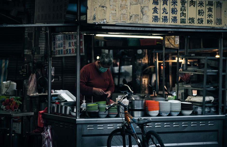 Masked street food vendor in an urban setting with colorful dishes and a bicycle nearby.