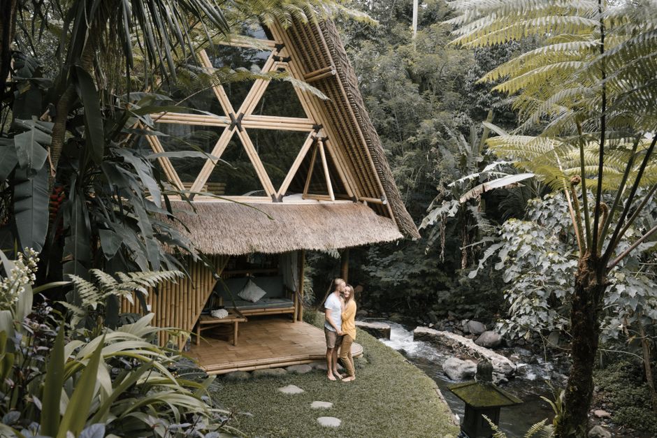 A couple embraces near a bamboo hut in a lush tropical garden by a gentle river.
