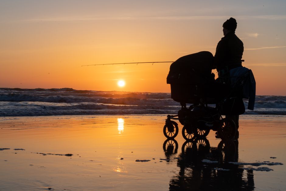 A serene scene of a parent pushing a stroller along a Danish shoreline, silhouetted against a vibrant sunset.
