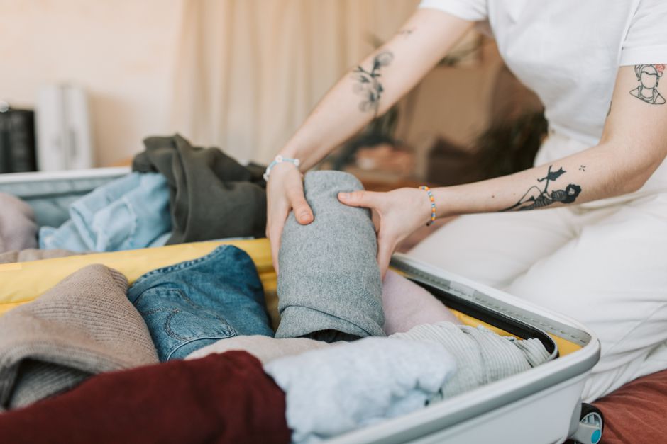 Close-up of a person packing clothes into a suitcase, showing arm tattoos, in a cozy indoo