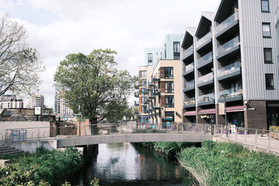 Contemporary apartment buildings line a calm river with a pedestrian bridge in the foregro