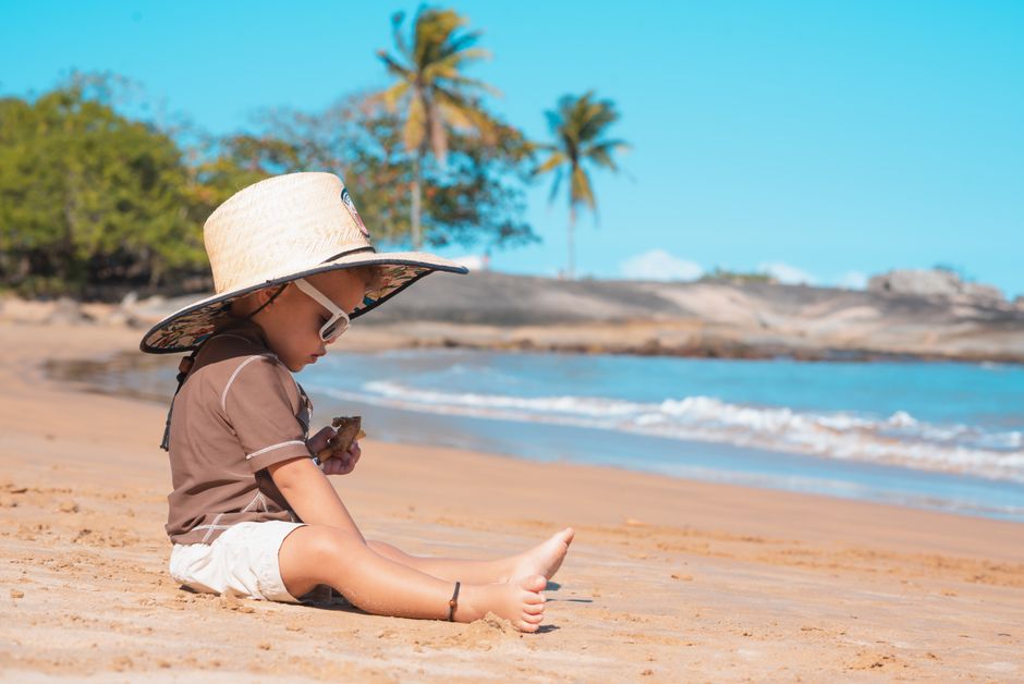 A young child wearing a sunhat sits on a sandy tropical beach playing with a toy, enjoying a sunny day.