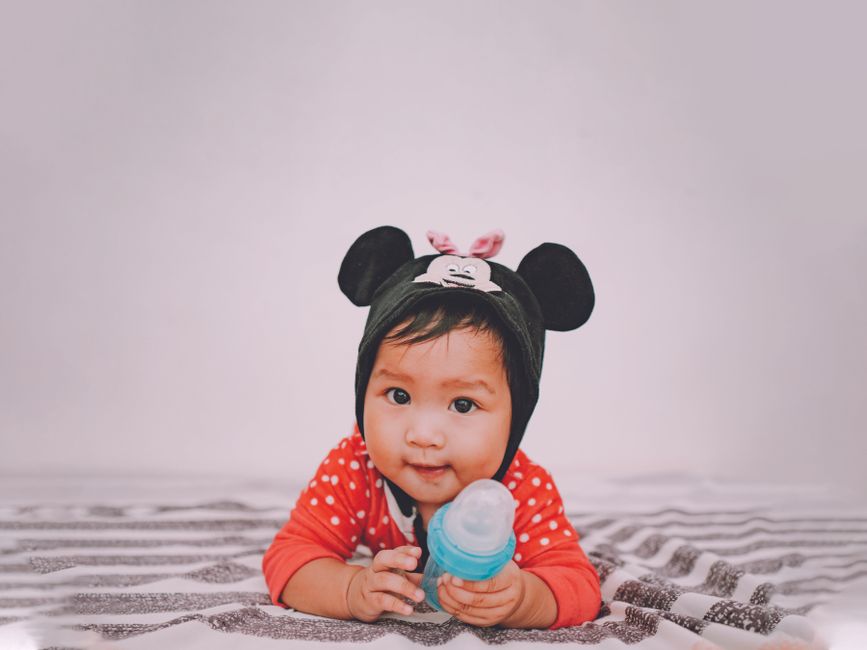 Cute baby wearing a mouse-themed hat, holding a bottle, and lying indoors on a striped blanket.