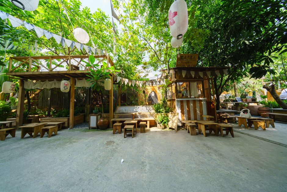 Charming outdoor cafe featuring wooden benches, lush greenery, and Japanese lanterns under a vivid blue sky.