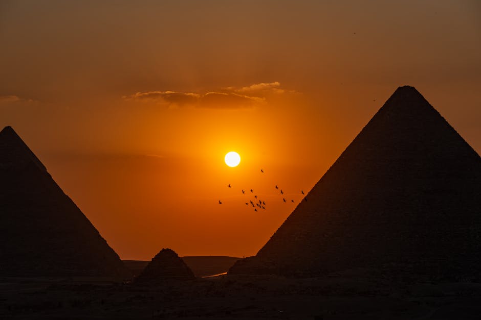 Sunset view of Egyptian pyramids with birds flying, creating a dramatic silhouette scene.