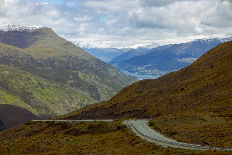 Winding road through mountains with a distant view of a lake and snowy peaks under a cloudy sky.
