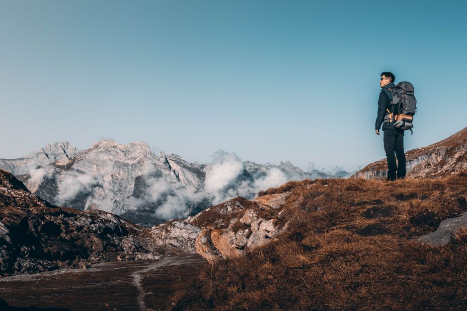 A hiker with a backpack stands on a rocky mountain trail, surrounded by stunning peaks and clouds under a clear blue sky.