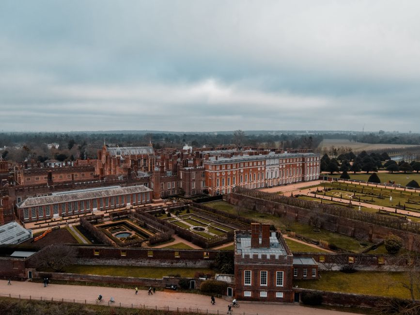 Drone view of Hampton Court Palace located in England in London with green grassy lawns with plants near fence and trees near pathways with people under cloudy gray sky
