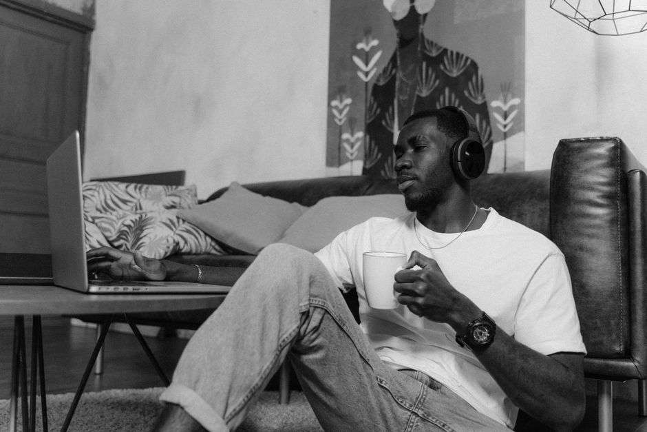 Black and white photo of a man relaxing with a coffee, headphones, and laptop in a living room.