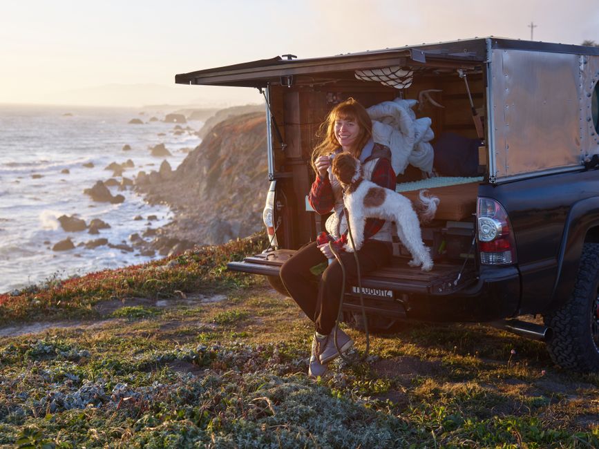 Woman and dog sitting in vehicle by ocean cliffs at sunset, exuding adventure.