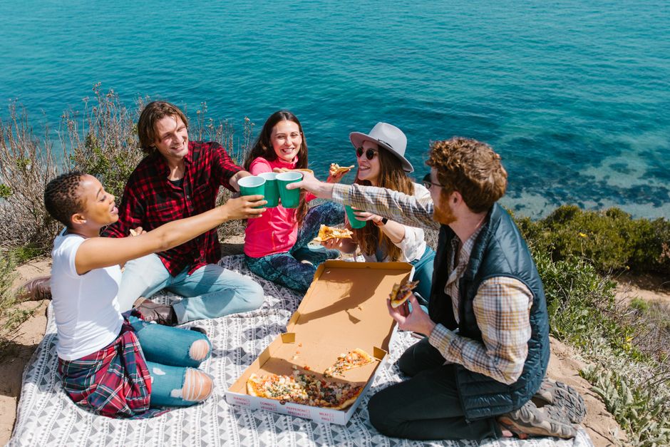 A diverse group of friends having a joyful picnic by the sea, enjoying pizza and drinks.