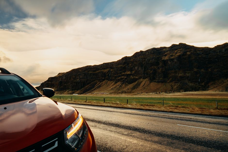 A red SUV travels along a rural road with a picturesque mountain landscape under a bright sky.
