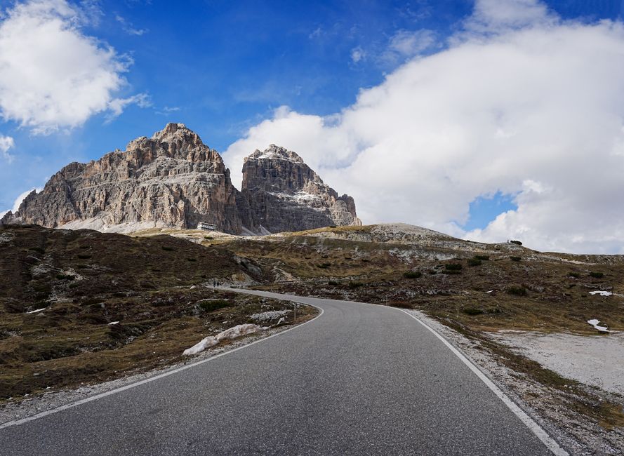 A winding road approaches the iconic Tre Cime di Lavaredo under a bright blue sky.