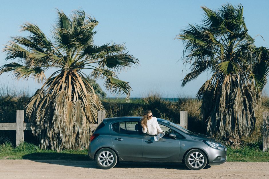Young woman in a car by palm trees on a scenic coastal road, embodying adventure and exploration.