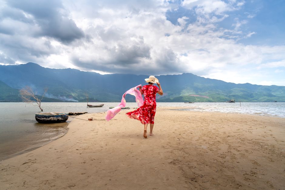 Back view full body of unrecognizable female traveler in dress and straw hat strolling on sandy beach near rippling sea against mountain ridge