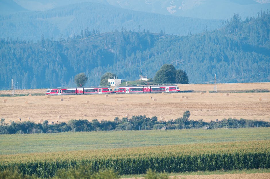 Red train travels through scenic fields in Poprad, Slovakia, surrounded by lush forests and mountains.