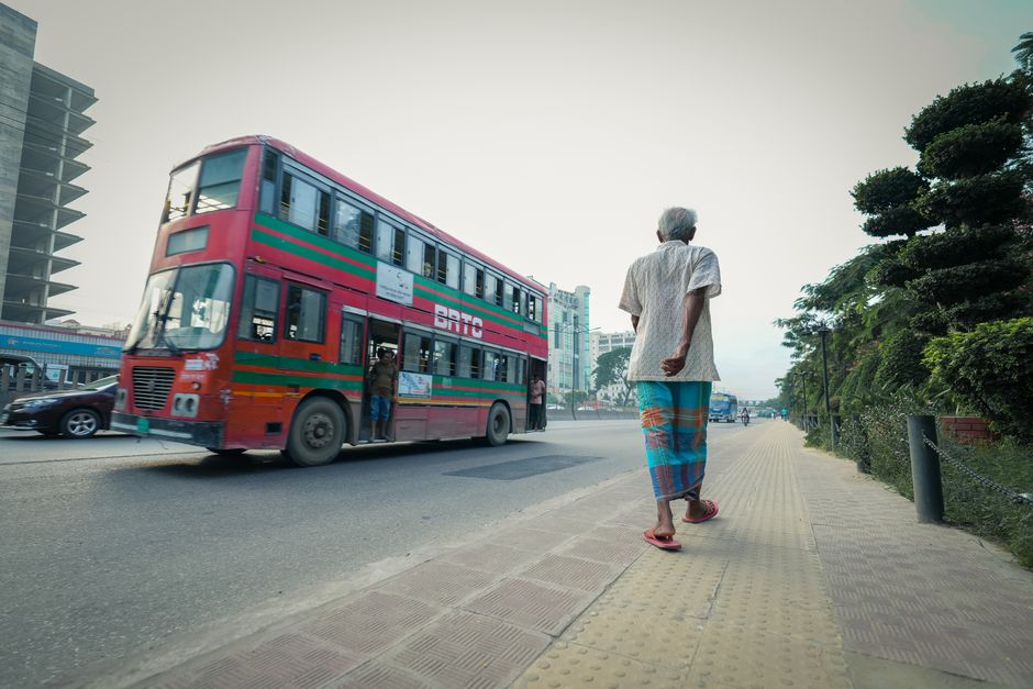 Elderly man walks along a busy city street with a vibrant double decker bus passing by on a sunny day.