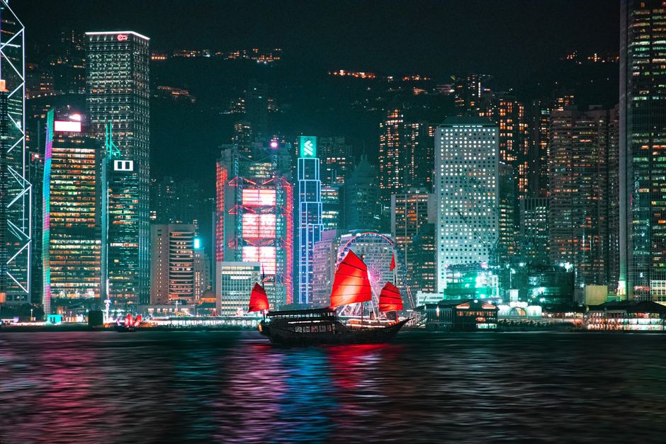 Vibrant Hong Kong skyline with a red-sailed junk boat on Victoria Harbour at night.