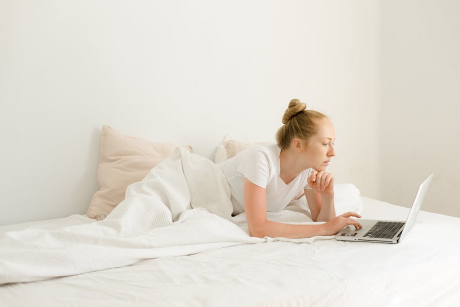 Young woman using a laptop while lying on a bed with a white blanket and pillows, enjoying a relaxed moment.