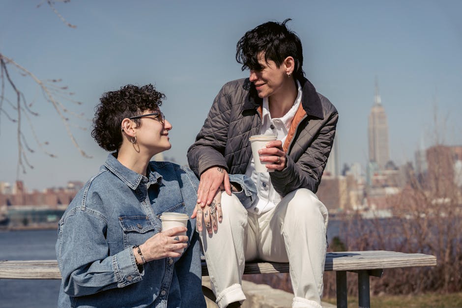 Enamored young lesbian couple with short dark hair in trendy outfits smiling and looking at each other while resting on wooden bench in city park and drinking takeaway coffee