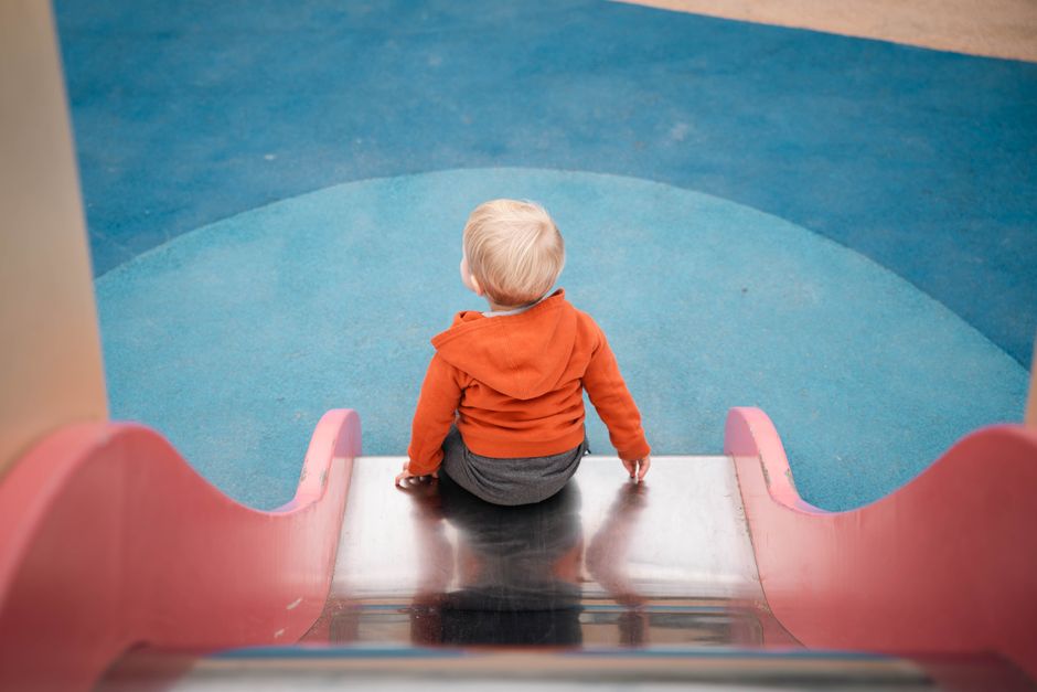 Young child in orange hoodie sits on a playground slide, ready to play.